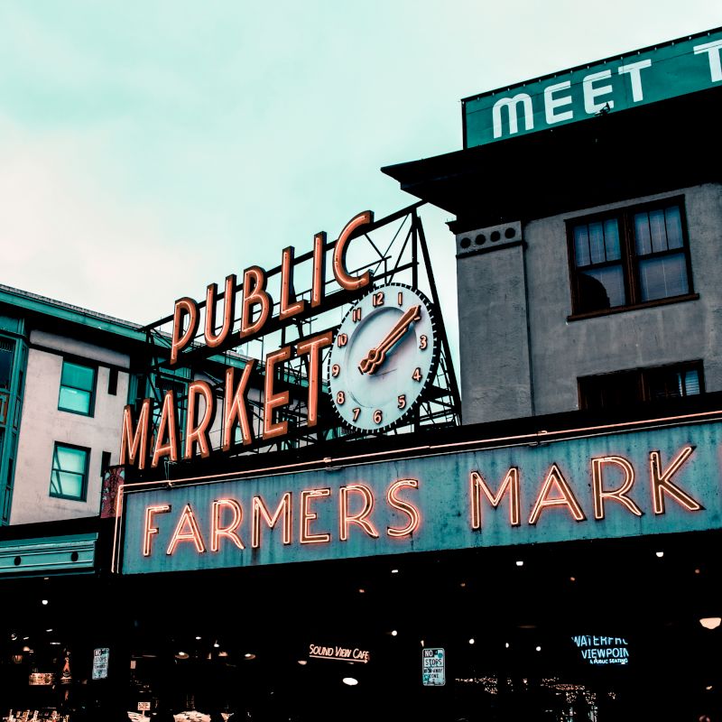 The image shows a neon sign for "Public Market" and "Farmers Market" with a clock, and nearby buildings, including one with a "Hotel" sign.
