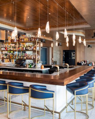A modern bar with a wooden countertop, blue barstools, pendant lights overhead, and a well-stocked shelf of various alcoholic beverages in the background.