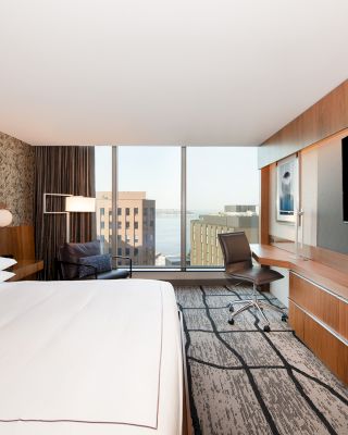 A modern hotel room features a large bed, a TV on a wooden unit, a desk with a chair, and a window with a city view.