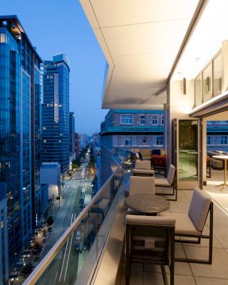A modern cityscape at dusk with tall buildings, seen from a balcony with tables and chairs, and large windows revealing an indoor area ending the sentence.