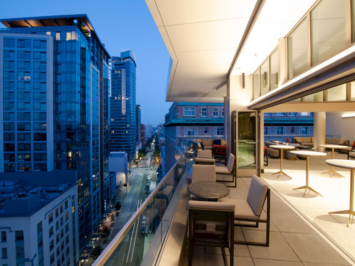 A modern rooftop terrace with seating areas overlooks an urban cityscape with tall buildings and streetlights illuminating the evening scene.