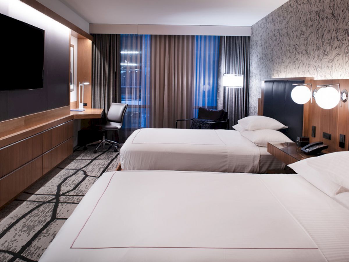 A modern hotel room with two beds, wall-mounted TV, desk with chair, and nightstand with lamps and phone, featuring contemporary decor.