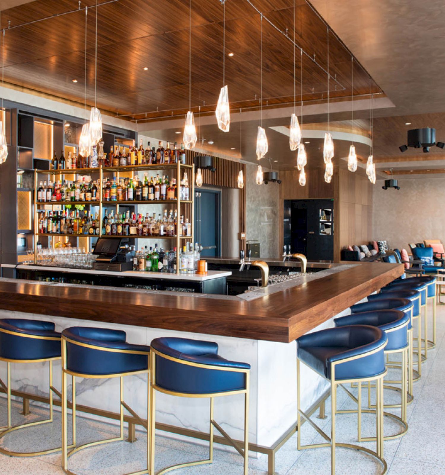A modern bar with a central counter, stylish hanging lights, and blue bar stools. Shelves behind the counter are stocked with various liquor bottles.