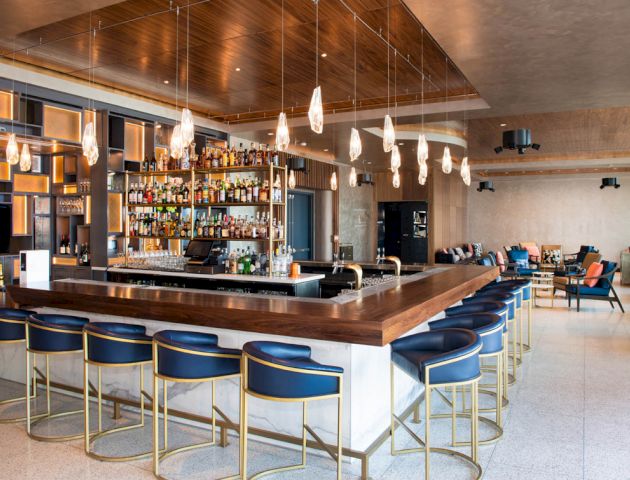 This image shows a modern bar with a well-stocked shelving unit behind it, stylish seating, pendant lights, and a lounge area in the background.