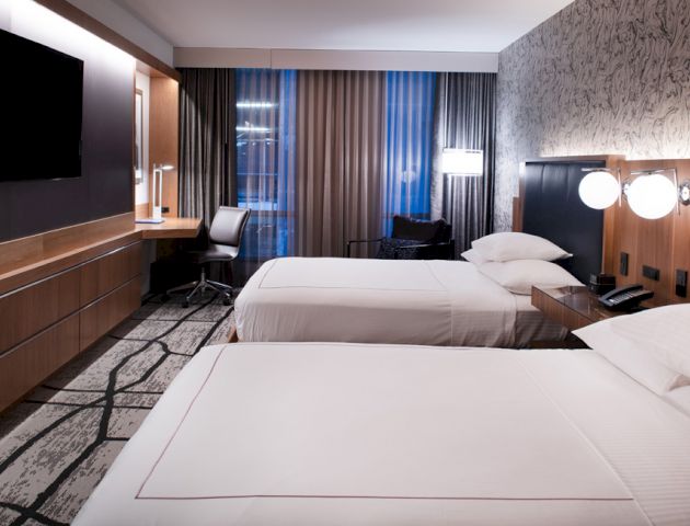 A modern hotel room with two neatly made beds, a wall-mounted TV, a desk with a chair, and a large window with curtains.