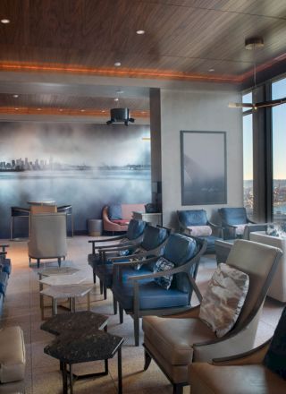 A modern lounge with numerous chairs, tables, a cityscape mural, and large windows offering a scenic view of a waterfront and Ferris wheel.