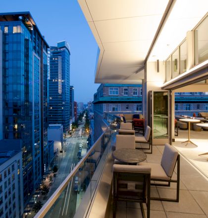 A modern balcony, overlooking a city street at dusk, with tables and chairs, and adjacent glass-walled rooms lit warmly from the inside.