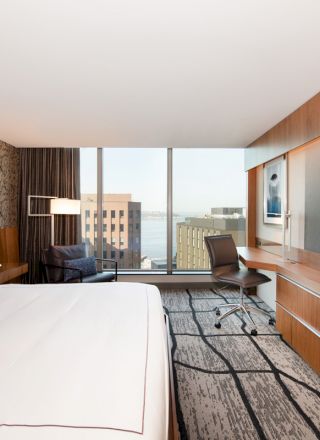 This image shows a modern hotel room with a large bed, a wall-mounted TV, a desk with a chair, and a view of buildings and water through the window.