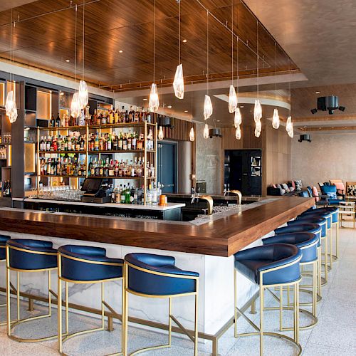 The image shows a modern bar with a wooden counter, blue bar stools, hanging lights, a stocked liquor shelf, and a lounge area with sofas.