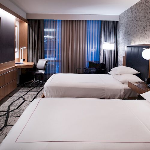 A modern hotel room with two neatly made beds, a TV mounted on the wall, a desk with a chair, and stylish decor.