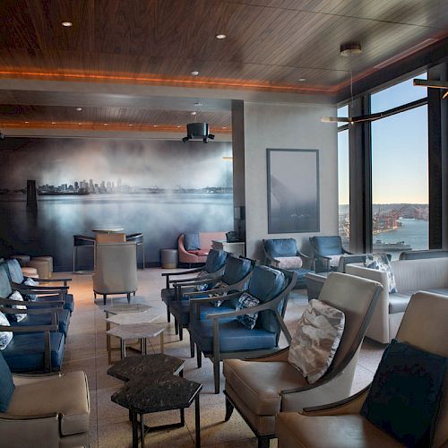 A modern lounge area with plush seating, large windows overlooking a waterfront cityscape, and a mural of a skyline on the back wall.