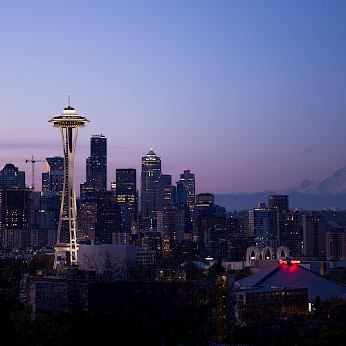 City skyline at dusk with prominent Space Needle tower, mountains in the background, and various buildings illuminated against a vibrant sky.