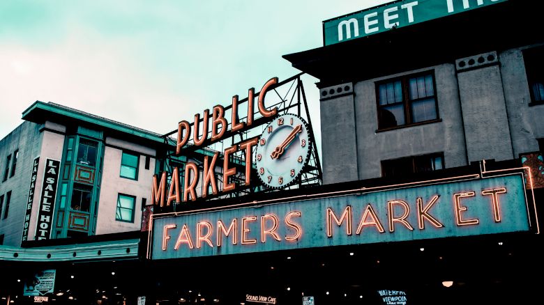 This image shows the iconic signage of the Public Market and Farmers Market, likely Pike Place Market, with surrounding buildings in the background.