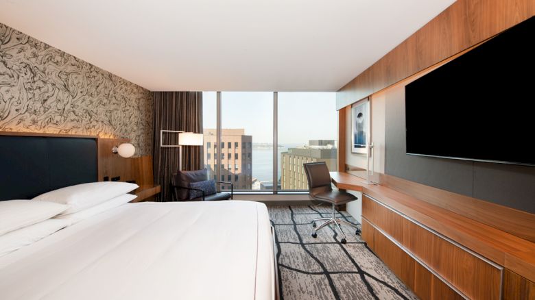 A modern hotel room with a large bed, wall-mounted TV, desk and chair, and a city view from floor-to-ceiling windows in the background.