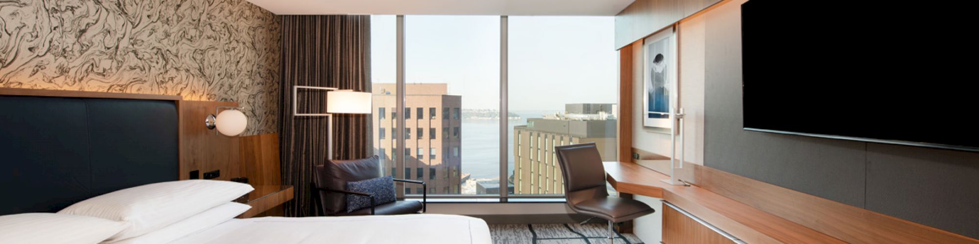 A modern hotel room featuring a large bed, desk with chair, flat-screen TV, and a window offering a city view with buildings and water.