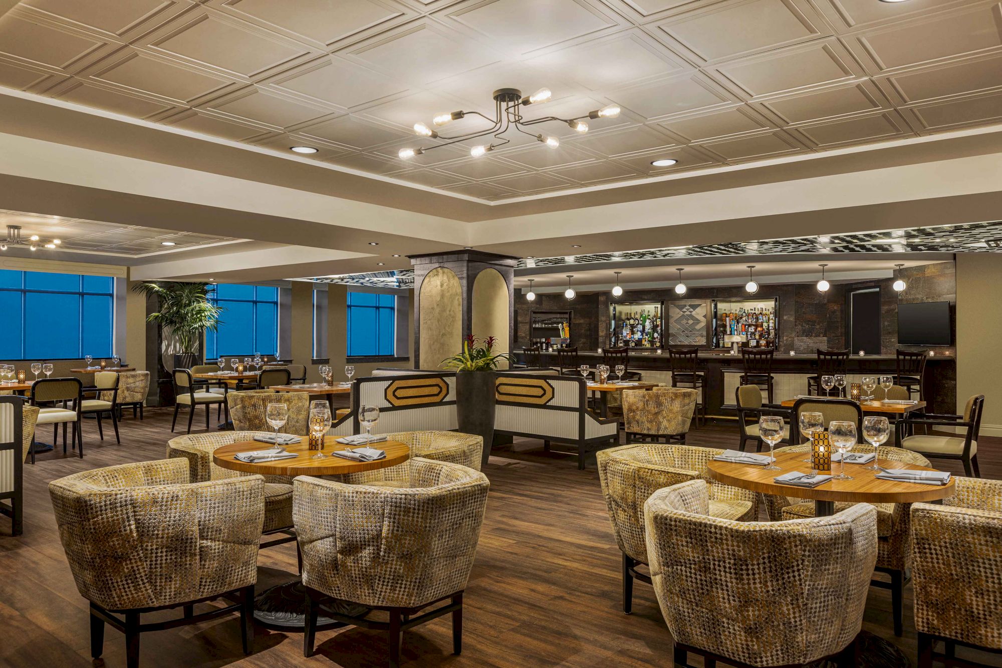 This image shows a modern, well-lit restaurant interior with circular tables, cushioned chairs, and a bar area in the background.