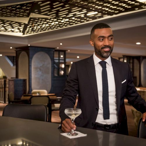 A well-dressed man stands at a bar holding a cocktail. The interior is stylish and modern, featuring ambient lighting and elegant decor.