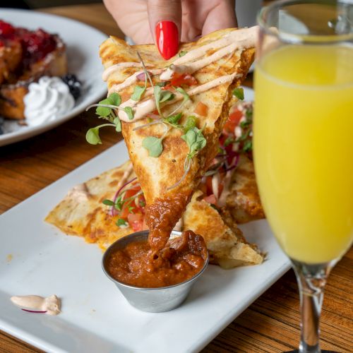 A hand with red nail polish dips a crispy quesadilla triangle into a red dipping sauce, surrounded by mimosas and other dishes on a wooden table.