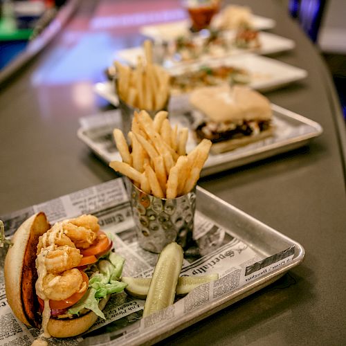 The image shows a line of trays with food, including burgers, fries, and a pickle spear, set along a counter in a restaurant or bar setting.