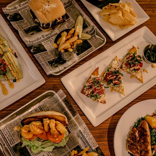 An assortment of dishes including a burger, fries, chips, tostadas with toppings, avocado slices, and a plate with meat and salad, all displayed on a wooden table.