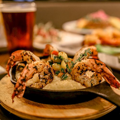 A dish with grilled shrimp atop a creamy base in a cast iron skillet, with a drink and other meals blurred in the background.