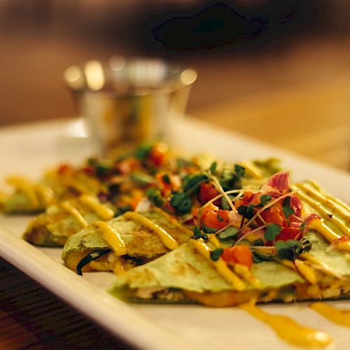 The image shows a plated dish of quesadillas topped with fresh salsa, herbs, and a drizzle of sauce, served on a long white plate with a dip.