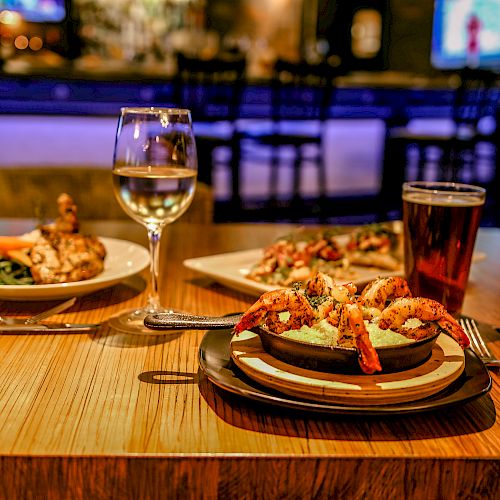 A restaurant table with shrimp pasta, chicken with vegetables, a glass of wine, a beer, and blurred background with bar and TV.