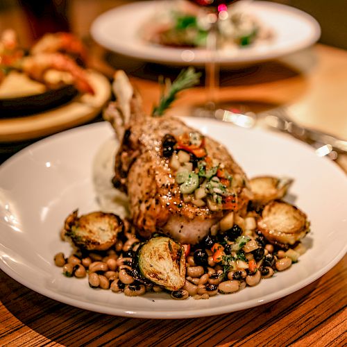 A gourmet plate featuring a seasoned chicken leg with beans and roasted vegetables, accompanied by a glass of red wine on a wooden table.