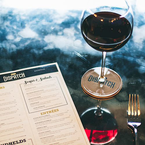 A glass of red wine is on a table beside a restaurant menu from 