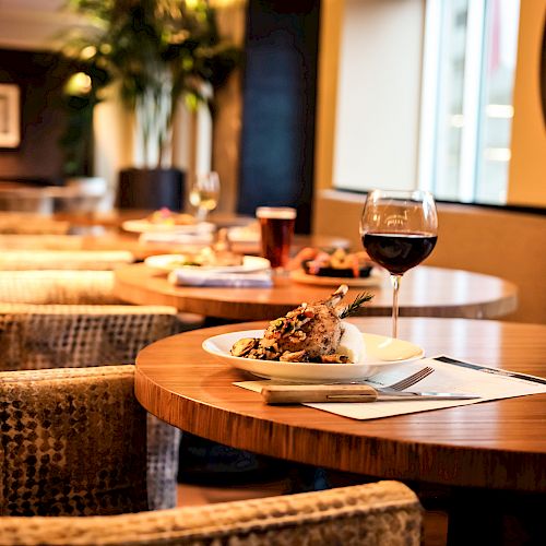 A restaurant table with a wine glass, a plated meal, and a notepad, surrounded by plush chairs in a dimly lit, elegant setting, is seen here.