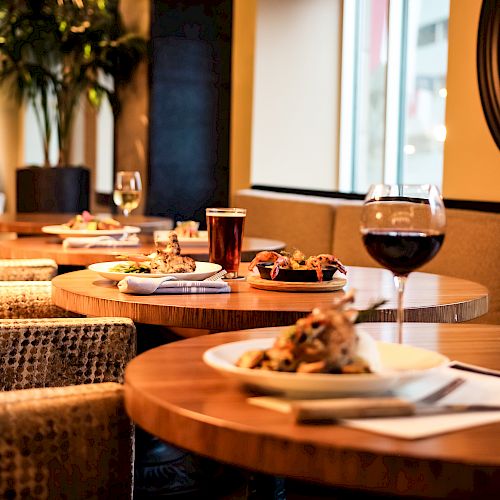 A cozy restaurant with wooden tables, plates of food, a glass of red wine, a glass of white wine, and a pint of beer on a table.