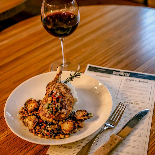 A plate of gourmet food and a glass of red wine on a wooden table, accompanied by a menu and cutlery, all set for a fine dining experience.