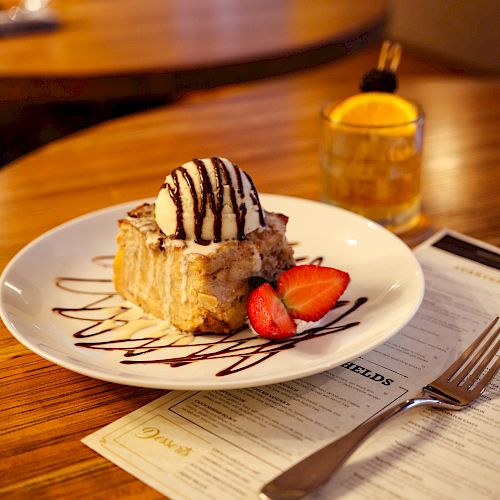 A dessert with ice cream and strawberries drizzled with chocolate sits on a plate on a wooden table beside a fork, a knife, and a glass of a drink.