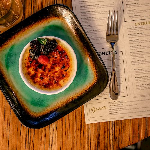 A brûlée dessert with berries sits on a plate next to a fork and a menu on a wooden table with a glass of drink.