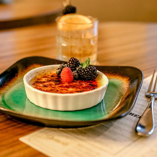 A crème brûlée topped with berries sits on a green plate next to a cocktail, a menu, and a fork on a wooden table in a cozy setting.