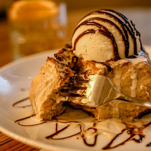 A dessert with ice cream and chocolate drizzle on a plate, with a fork in it and a strawberry garnish. A drink is blurred in the background.