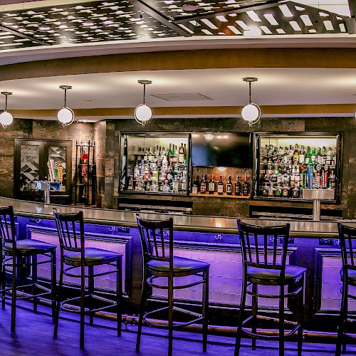 This image shows an empty bar with high chairs, a stocked liquor shelf, modern lighting, and a stylish ceiling design.