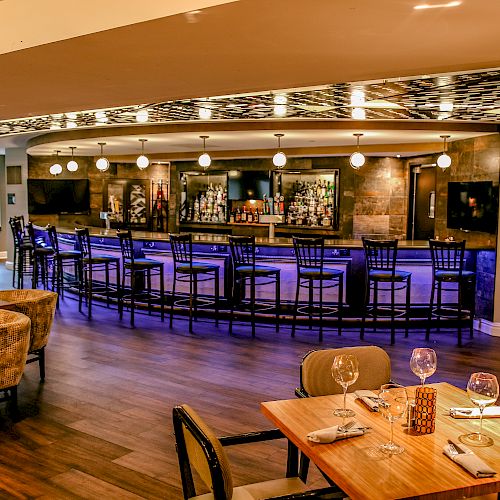 The image shows a modern, well-lit bar with bar stools, a stylish dining area with set tables, and comfortable seating.