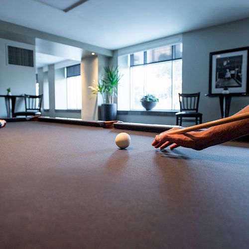 A person is playing pool on a billiard table inside a room with large windows, chairs, and a framed picture on the wall. The cue ball is in focus.