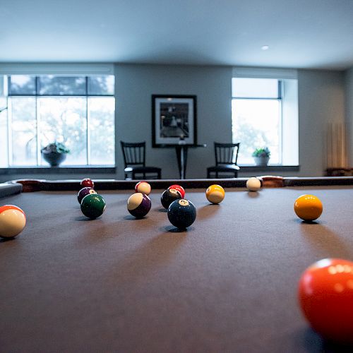 A billiard table with balls scattered across it in a room featuring large windows, chairs, and potted plants.