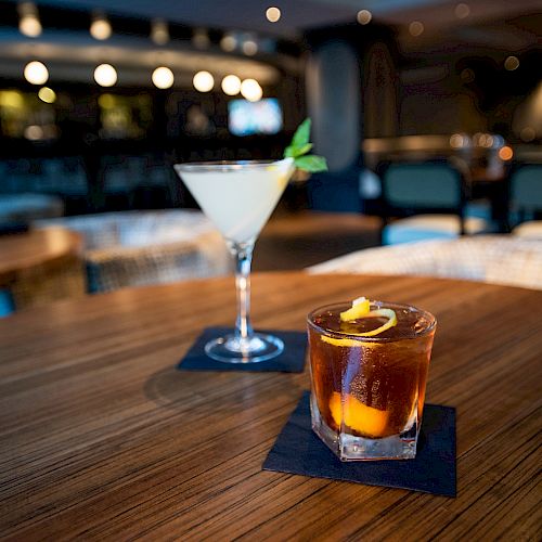 A martini and an old-fashioned cocktail on a wooden table in a dimly lit bar or restaurant setting with chairs and tables in the background.