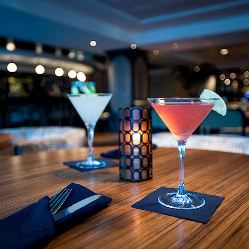 This image shows two cocktails on a bar table, a candle in the center, and a dimly lit bar in the background.