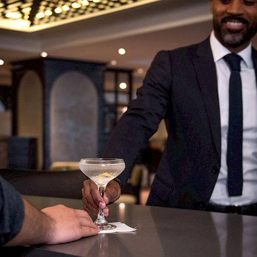 A man in a suit hands a martini glass to another person, standing at a polished bar counter in an upscale setting.