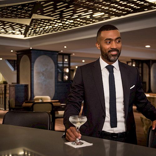 A well-dressed man in a suit stands at a modern bar holding a martini glass, with an elegant interior featuring ambient lights and stylish decor.