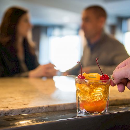 A hand holding a cocktail garnished with cherries and an orange slice, with three people conversing in the background at a bar counter.