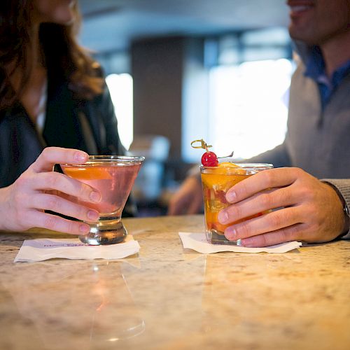 Two people holding cocktails, sitting at a bar, and partially facing each other. The drinks are on napkins atop the countertop, with blurred backgrounds.