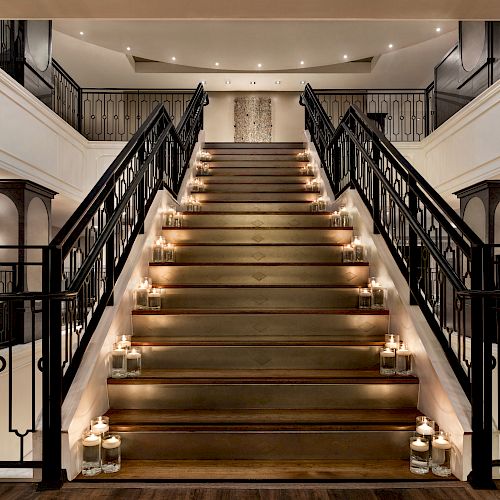 A grand staircase with black railings, lined with candles and leading to an upper level in a well-lit, elegant interior space.