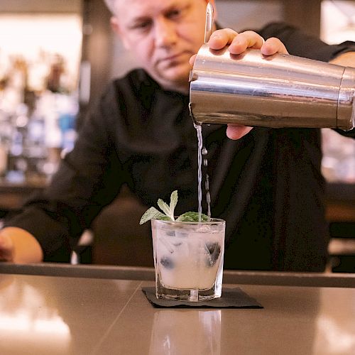 A bartender is pouring a drink from a shaker into a glass with mint leaves at a bar.