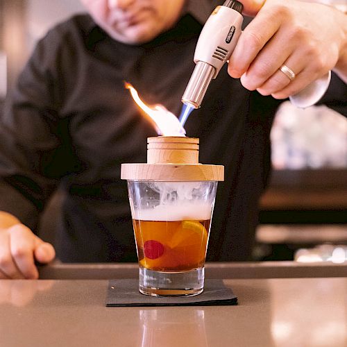 A person is using a culinary torch to heat a wooden cover on top of a drink, possibly for smoking or flavor enhancement purposes.