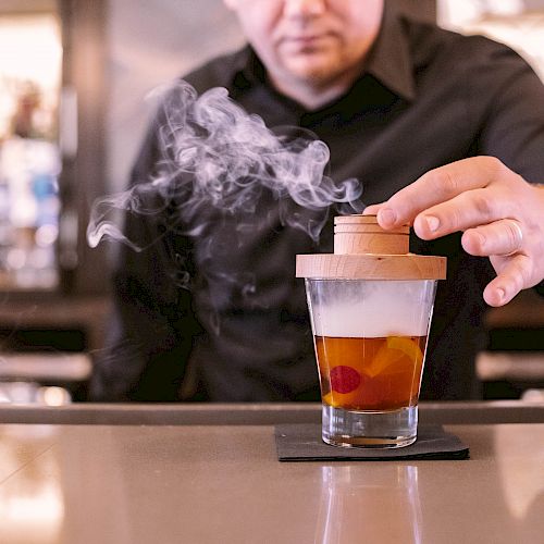 A person places a wooden lid on a glass filled with a beverage, while smoke emerges from the drink, creating a visually appealing effect.